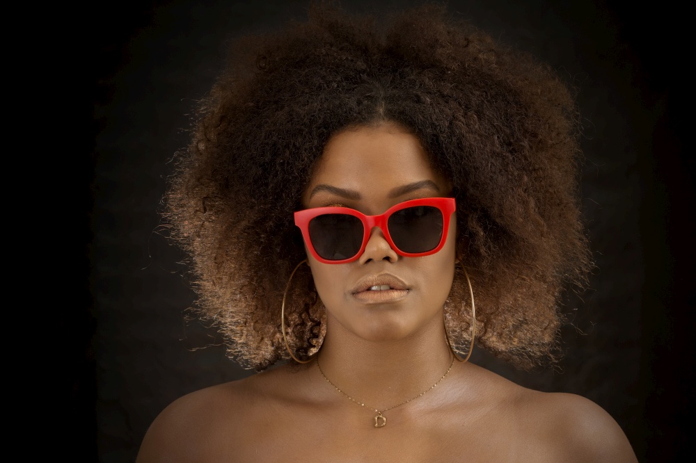 Modern black female model with Afro hairstyle and bare shoulders wearing sunglasses in dark studio. Cool African American woman in red sunglasses