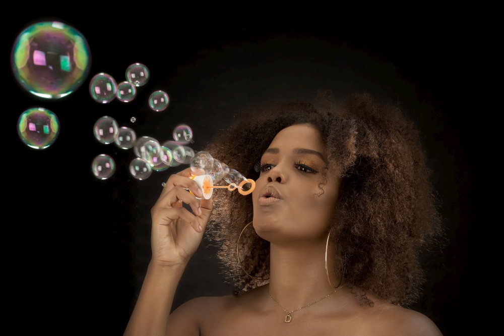Flirty young ethnic female with Afro hairstyle blowing soap bubbles on black background. Charming black woman blowing soap bubbles in studio