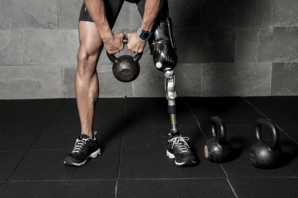 Faceless shot of sportsman with artificial leg limb training with kettlebell in gym. Crop man with prosthesis lifting kettlebell