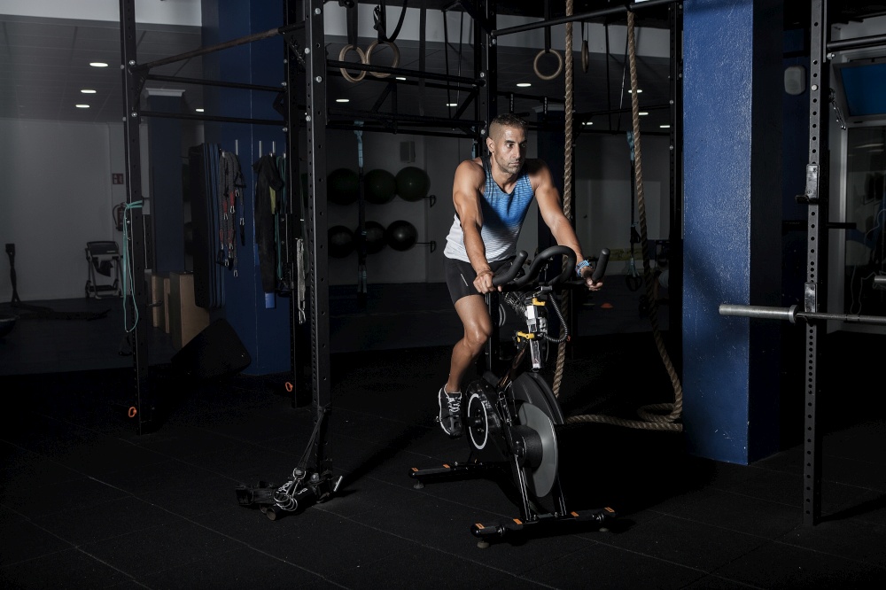 Adult confident sportsman riding stationary bicycle in modern dark gym. Sportsman riding stationary bicycle in gym