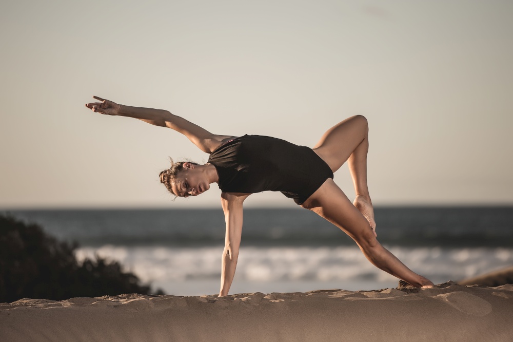 Beautiful slim woman in black bodysuit training yoga pose on sandy beach with ocean landscape. Graceful woman doing asana on beach