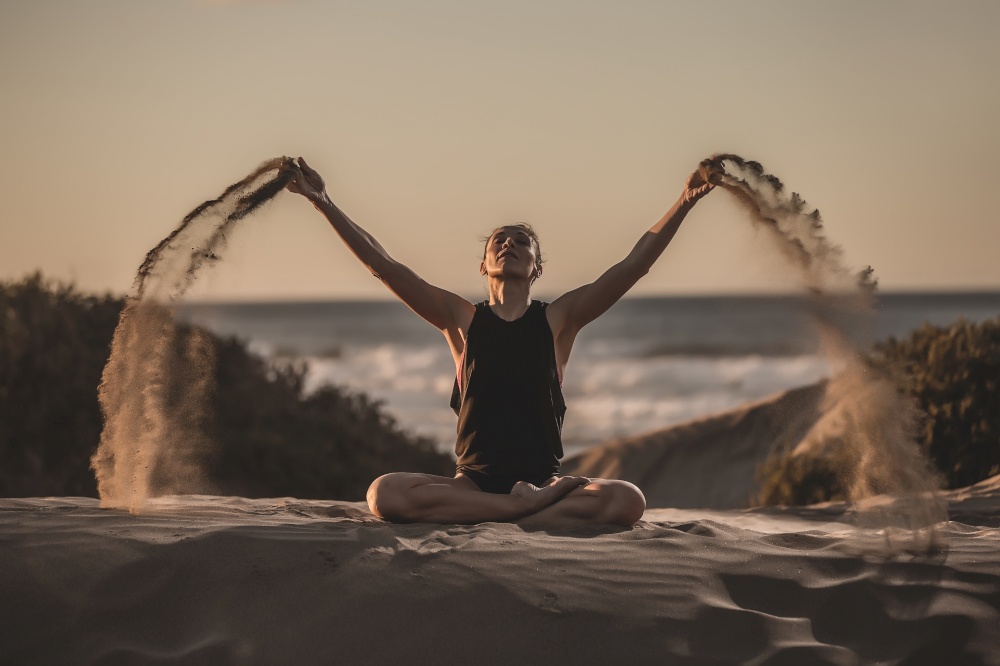 Sensual woman sitting in meditation pose while pouring heaps of sand in sunlight. Woman on seashore in asana with flying sand