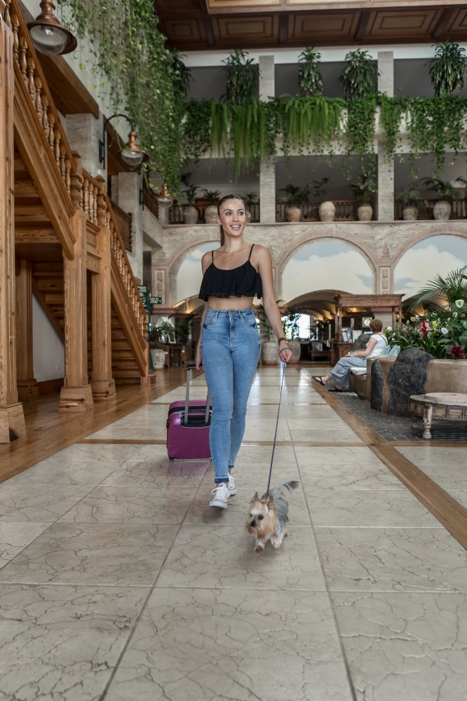 Woman with suitcase and Yorkshire Terrier walking in lobby of luxury hotel during vacation. Female traveler with dog walking in hotel