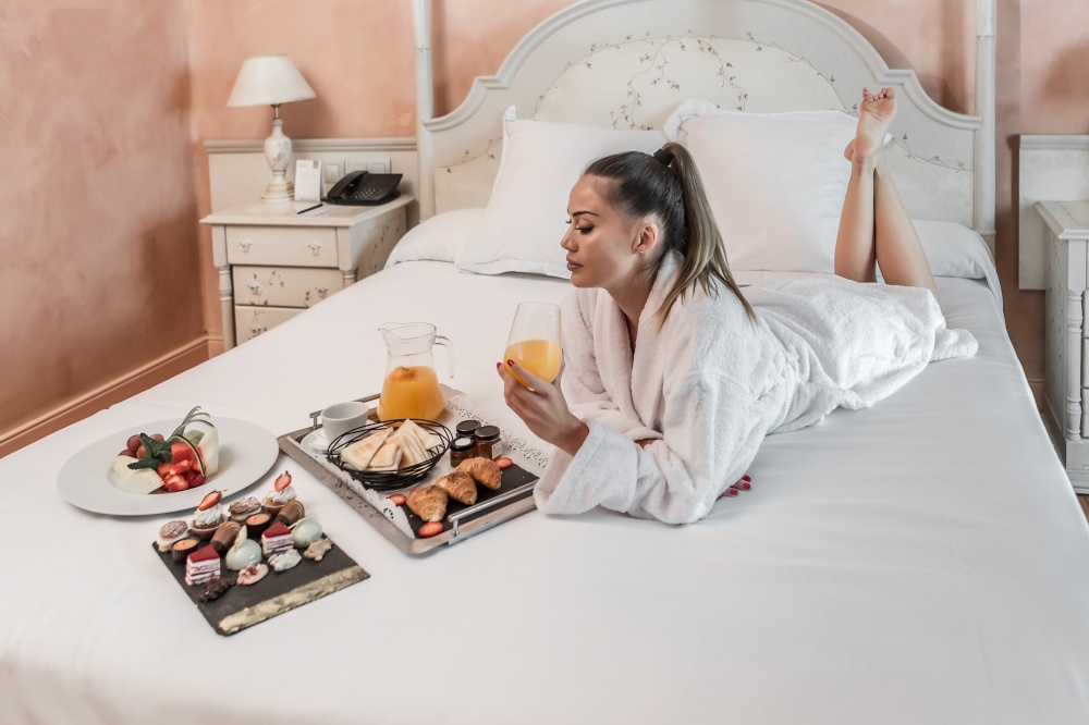 Attractive barefoot female in bathrobe holding glass of fresh juice and looking at various breakfast food while lying on bed in hotel room. Barefoot woman enjoying breakfast on bed