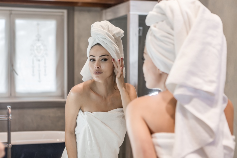 Beautiful female in white towels smearing cream on clean face skin while looking at mirror in bathroom after shower. Woman applying cream after shower