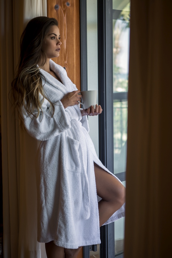 Side view of attractive female with loose long hair in bathrobe holding cup in hands standing at open balcony and looking away. Young woman in bathrobe standing at window