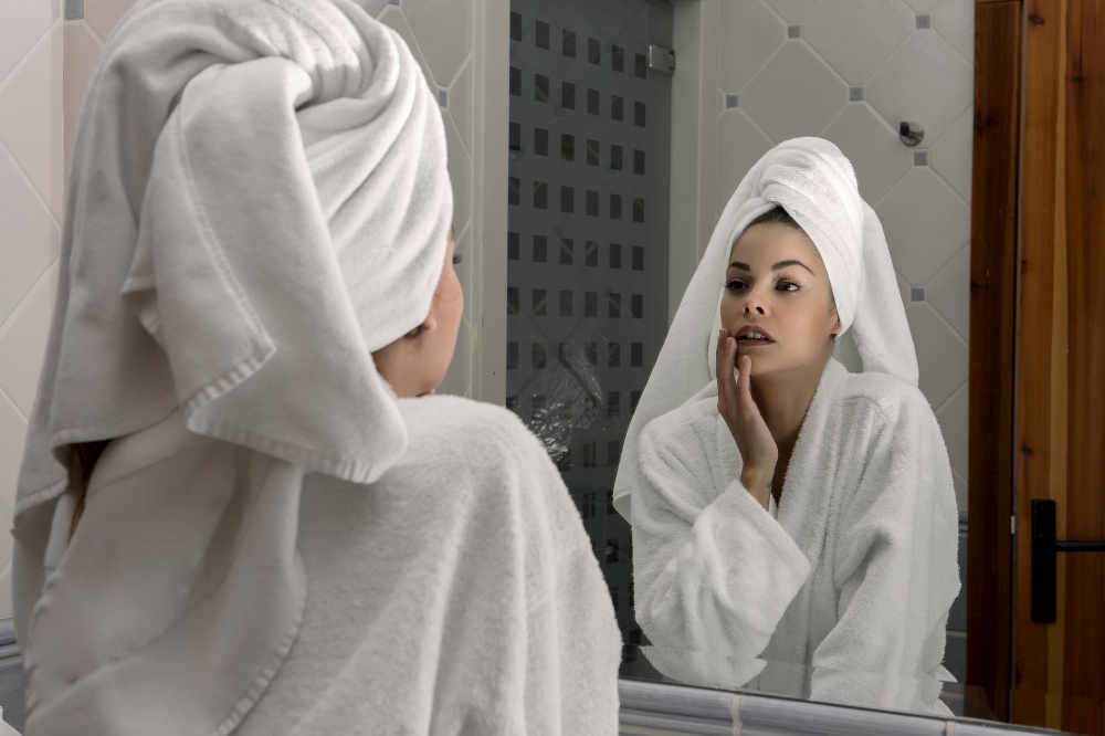 Sensual young woman in white bathrobe and with towel on head touching gentle skin looking at mirror in bathroom. Alluring woman in towel touching face