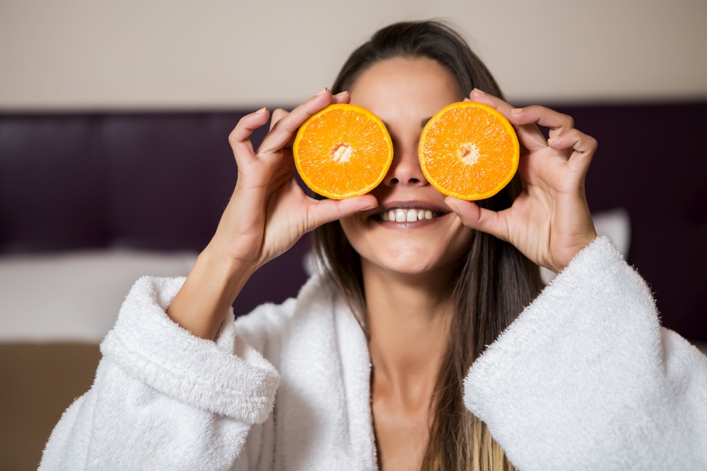 Pretty young brunette woman in white morning gown sitting in hotel room and holding slices of orange near eyes. Woman putting slices of orange near eyes