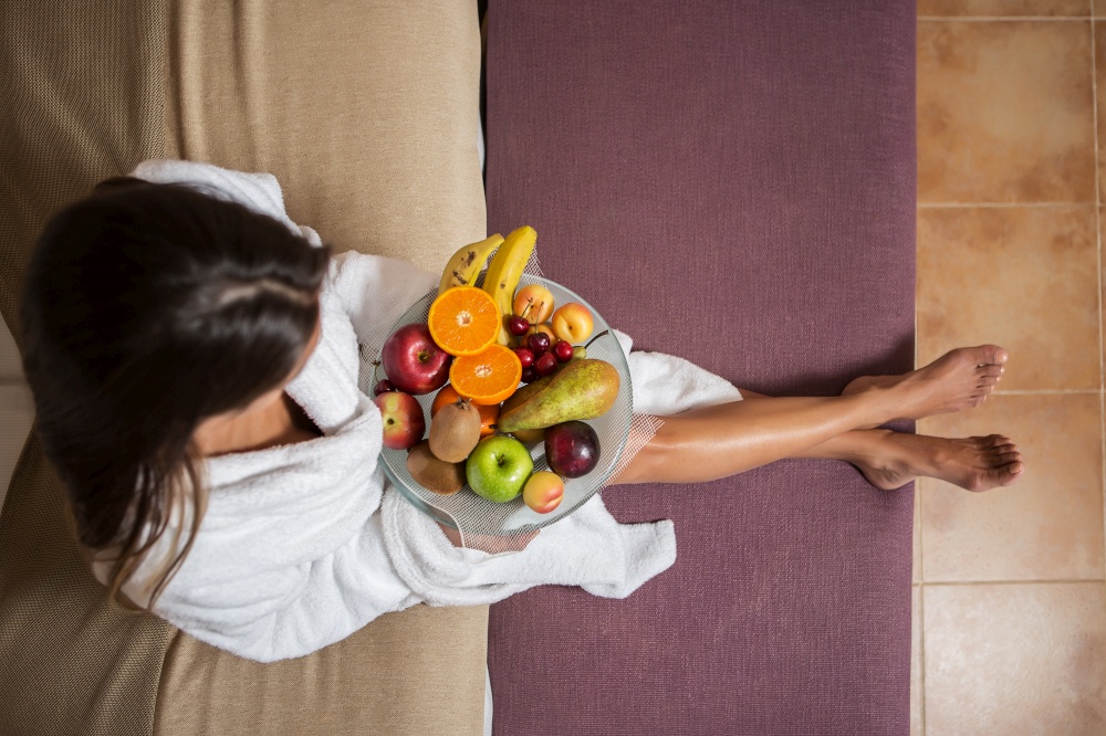 From above shot of woman in white bathrobe sitting on bed with plate of colorful fruit . Woman with fruit mix on bed