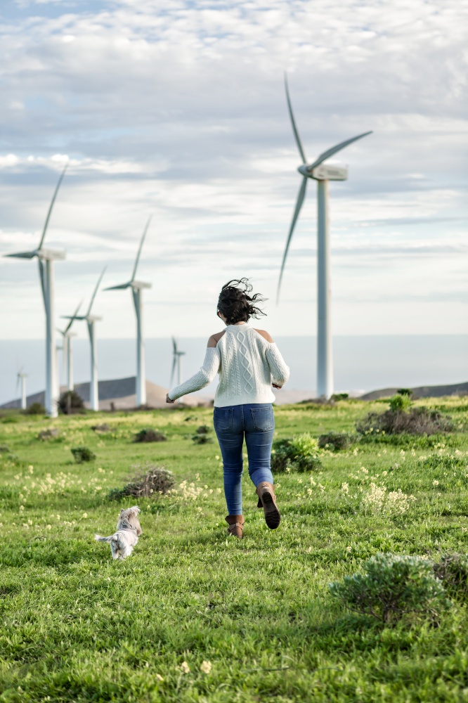 Back view of unrecognizable female running along blooming meadow with little dog while enjoying summer weekend against windmills on Lanzarote. Woman running along field with dog in summer