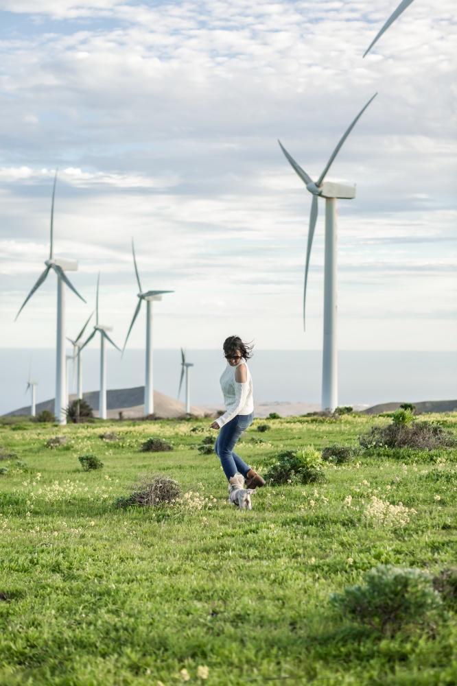 Side view of carefree female running in field with windmills and playing with adorable dog while enjoying summer weekend on Lanzarote. Woman playing with dog in meadow with windmills