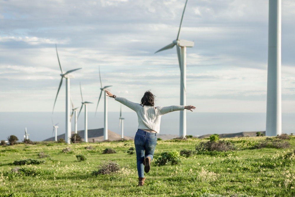 Back view of carefree female running with outstretched arms in field with windmills on sunny day in summer on Lanzarote. Woman running in meadow in summer