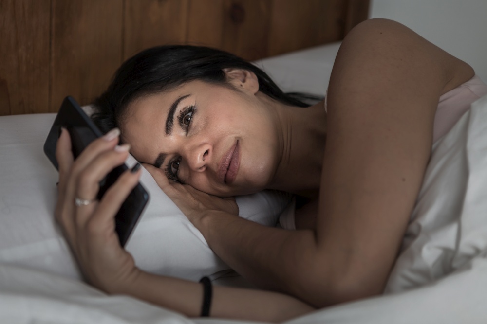 Delighted female in pajama lying under blanket in bed and surfing Internet via mobile phone before sleep. Content woman lying in bed and browsing smartphone