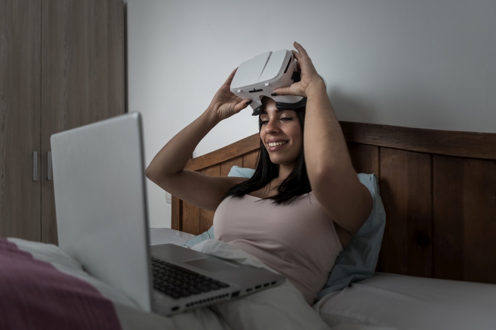 Content female putting on glasses of virtual reality while sitting on bed with laptop and preparing for experiencing cyberspace at home. Woman using VR goggles and laptop on bed