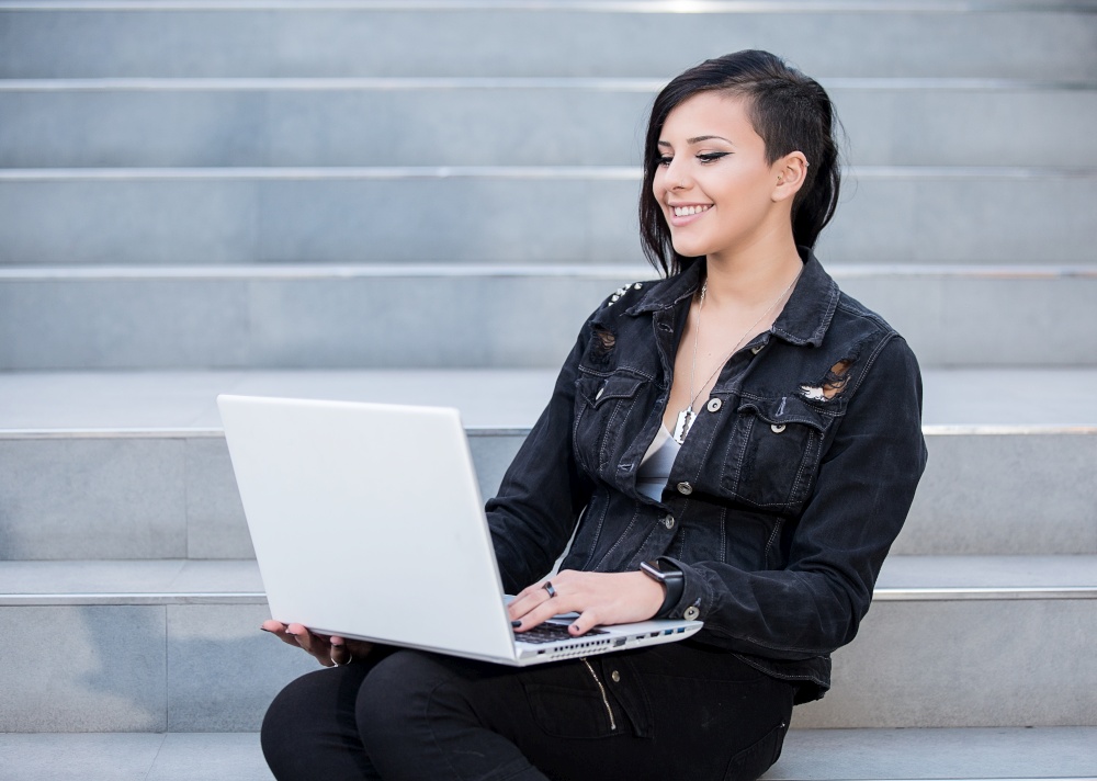 Smiling casual dressed woman sitting on steps and using laptop.. Woman with laptop on stairs