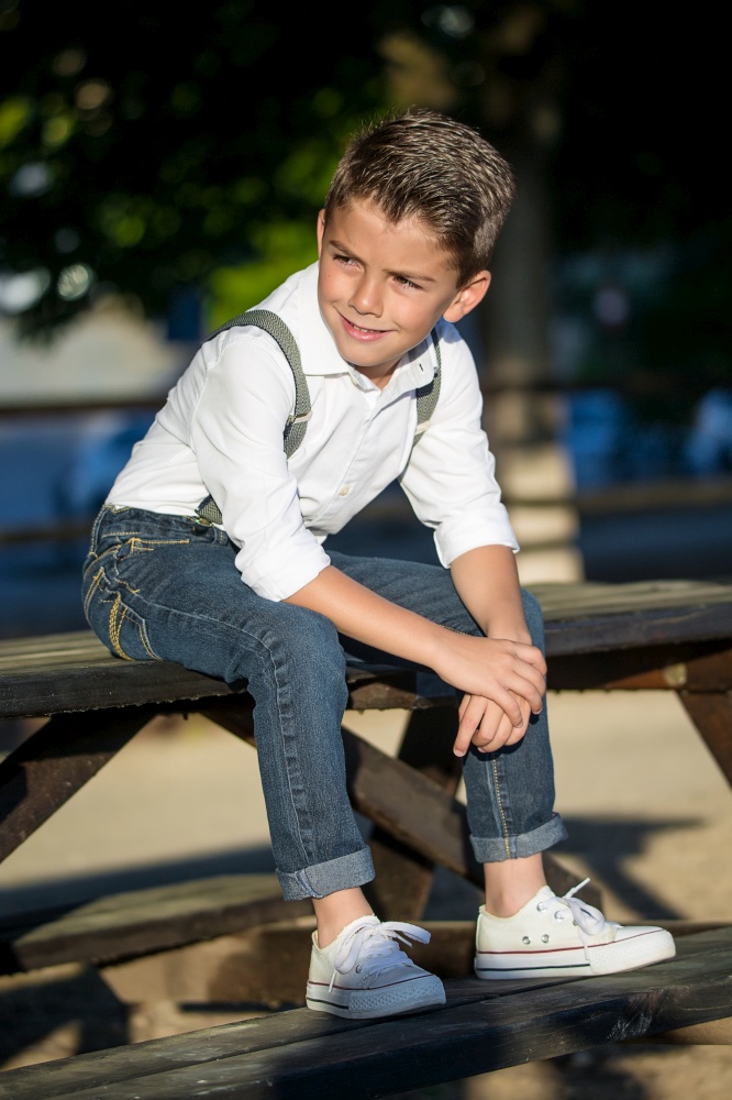 Stylish little boy in white shirt and suspenders posing in park and looking away.. Dandy kid posing in sunlight