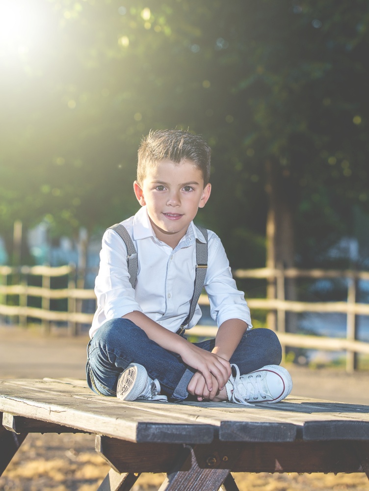 Stylish boy in suspenders and jeans sitting on table in park looking at camera.. Adorable trendy kid posing in park