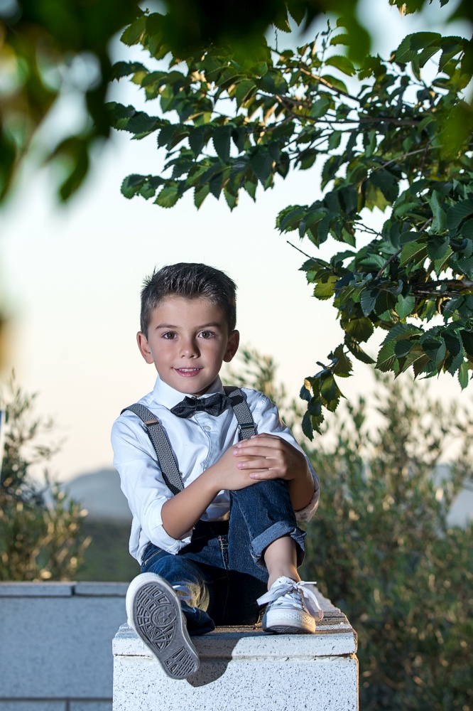 Adorable little boy wearing stylish outfit posing confidently at camera sitting on fence.. Wonderful little dandy posing at camera