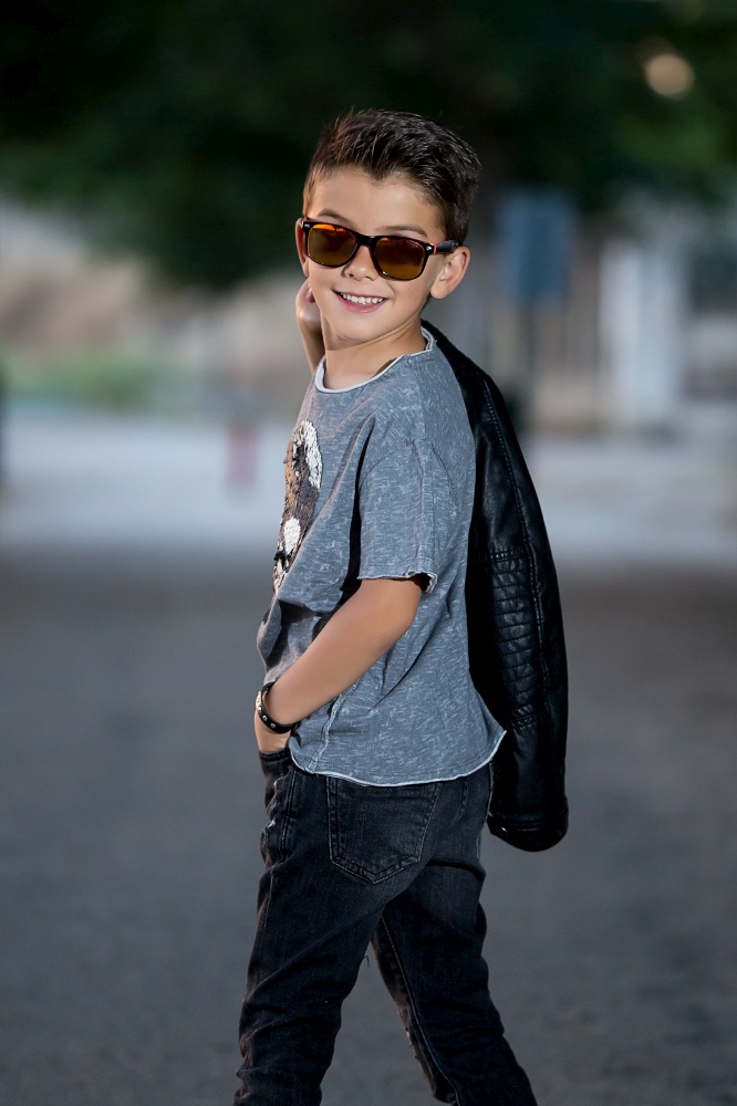 Back view of stylish rocker in sunglasses holding jacket on shoulder and smiling over shoulder.. Charming trendy boy posing at camera