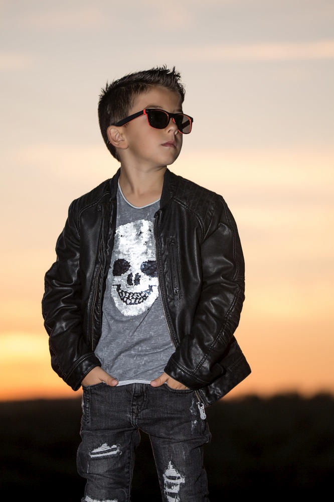 Stylish boy wearing sunglasses and black leather jacket standing with hands in pockets on nature.. Confident rocker kid in sunset