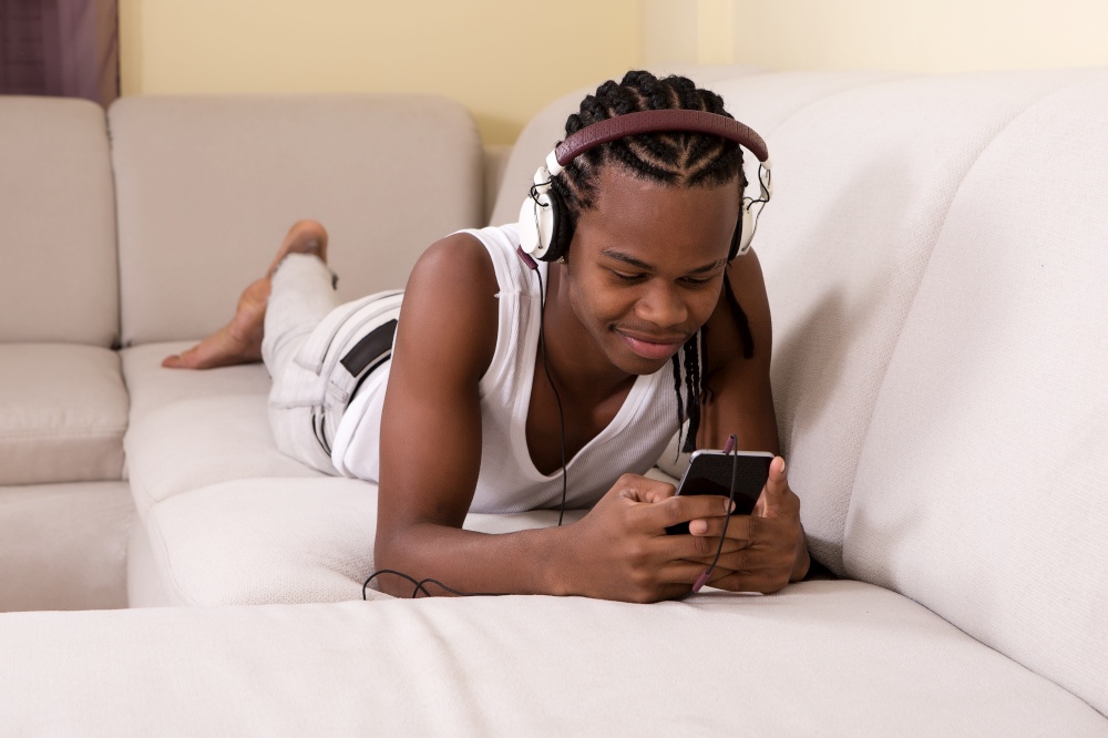 Young black man in headphones lying and relaxing on couch with smartphone.. Young man lying on sofa with phone