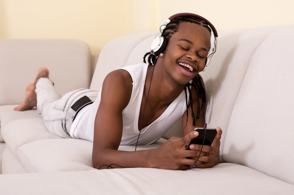 Cheerful black man in headphones lying on sofa and using smartphone.. Smiling man lying and using phone