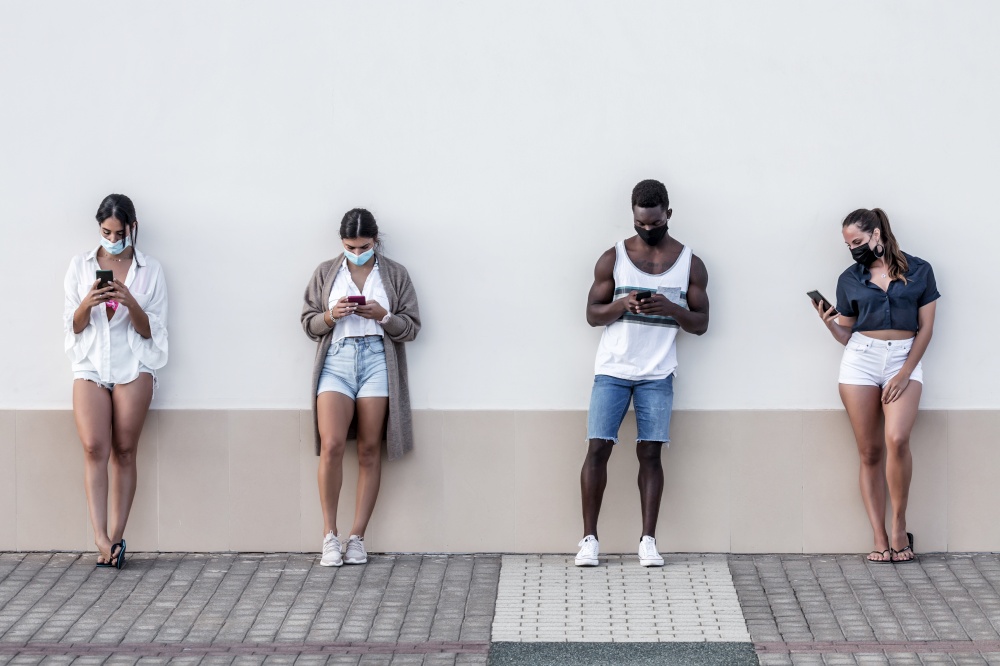 Group of multiethnic friends in masks standing near urban building and surfing Internet on mobile phones on Lanzarote. Diverse friends browsing smartphones on street
