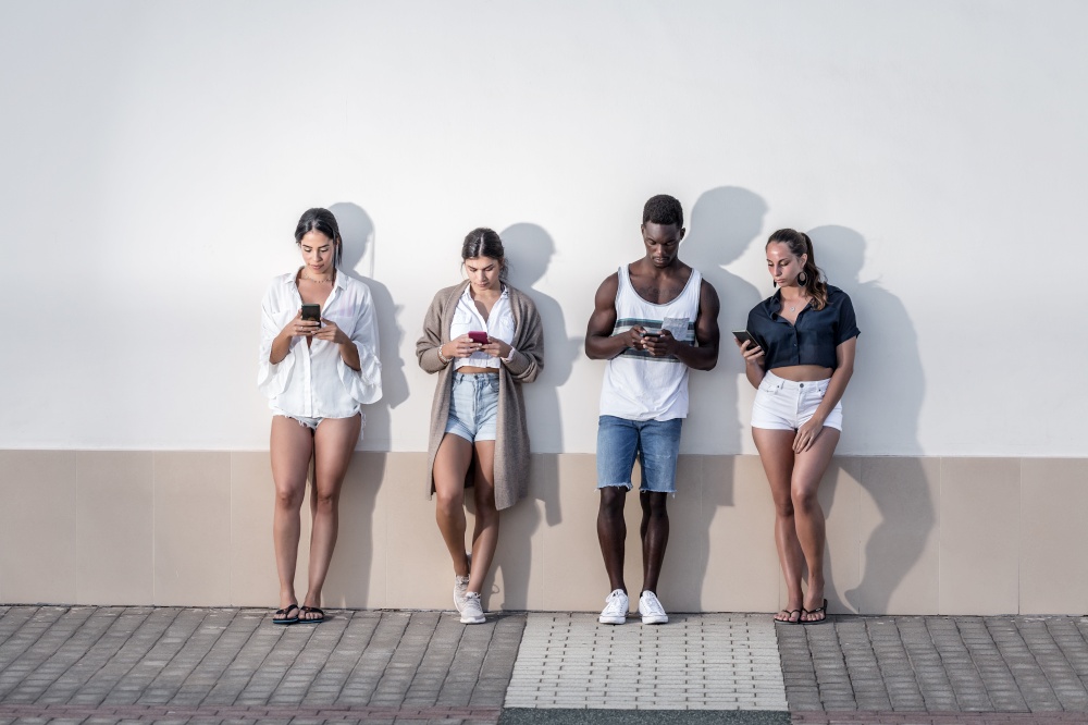 Company of multiethnic friends standing near building in city and browsing mobile phones while spending weekend on Lanzarote. Group of diverse people using smartphones on city street