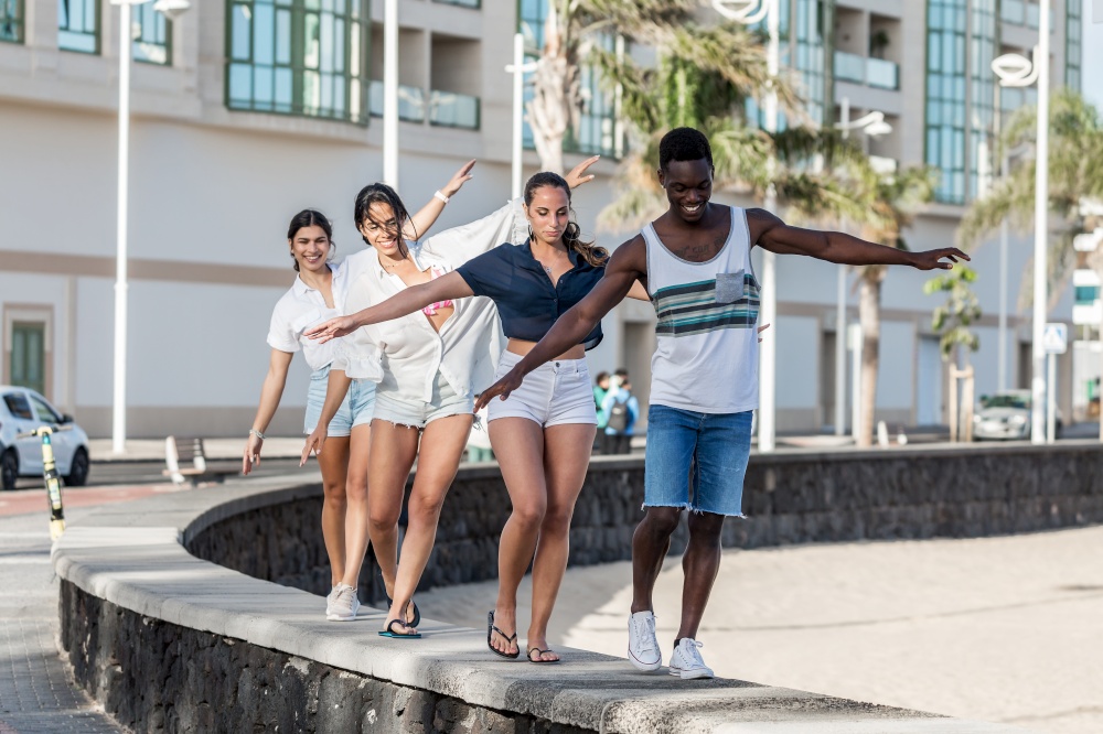 Positive diverse friends walking along stone fence and pretending flying airplane while having fun at weekend on Lanzarote. Multiethnic friends having fun together in city