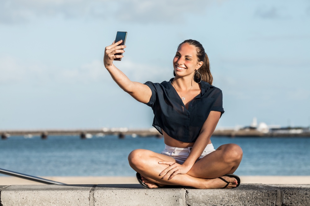 Glad female sitting on stone fence on seashore and taking self shot on smartphone while enjoying weekend on Lanzarote in summer. Smiling woman taking selfie on embankment