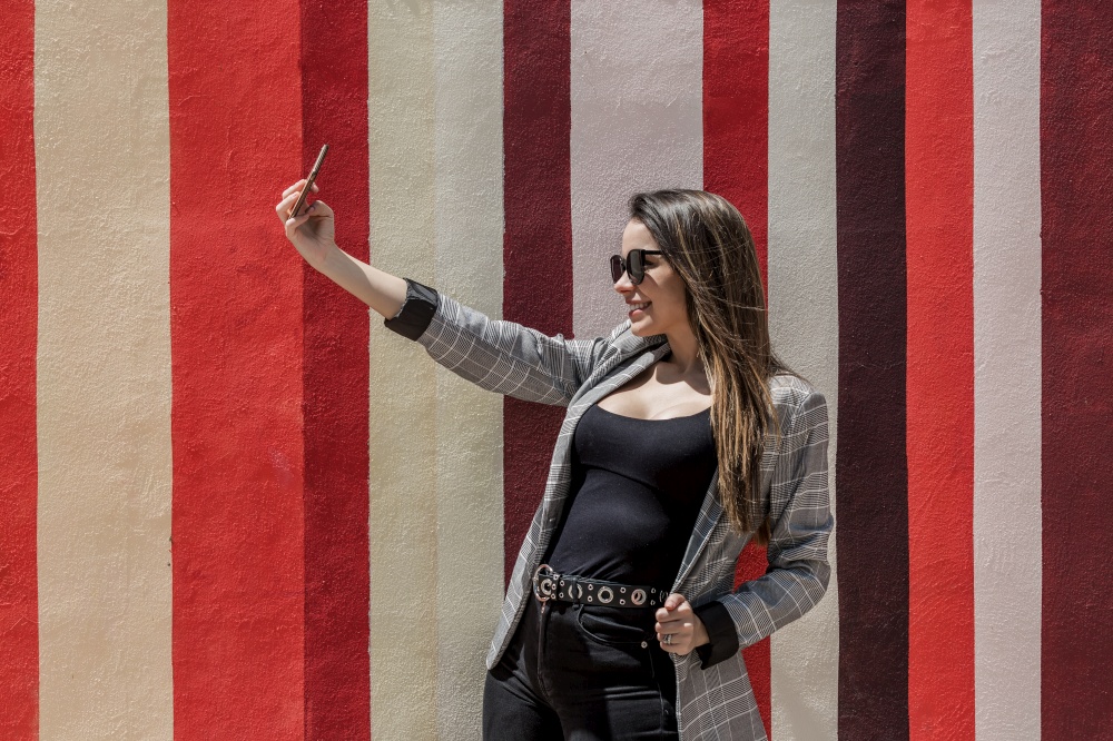 Content female in trendy outfit taking self portrait on mobile phone in street against colorful striped background. Stylish woman taking selfie in city on sunny day