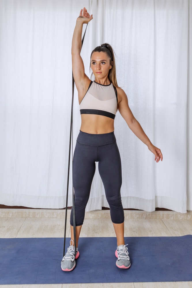 Full body of active fit young female in sportswear doing single arm shoulder press exercise with resistance band during fitness workout at home. Fit woman doing resistance band workout at home