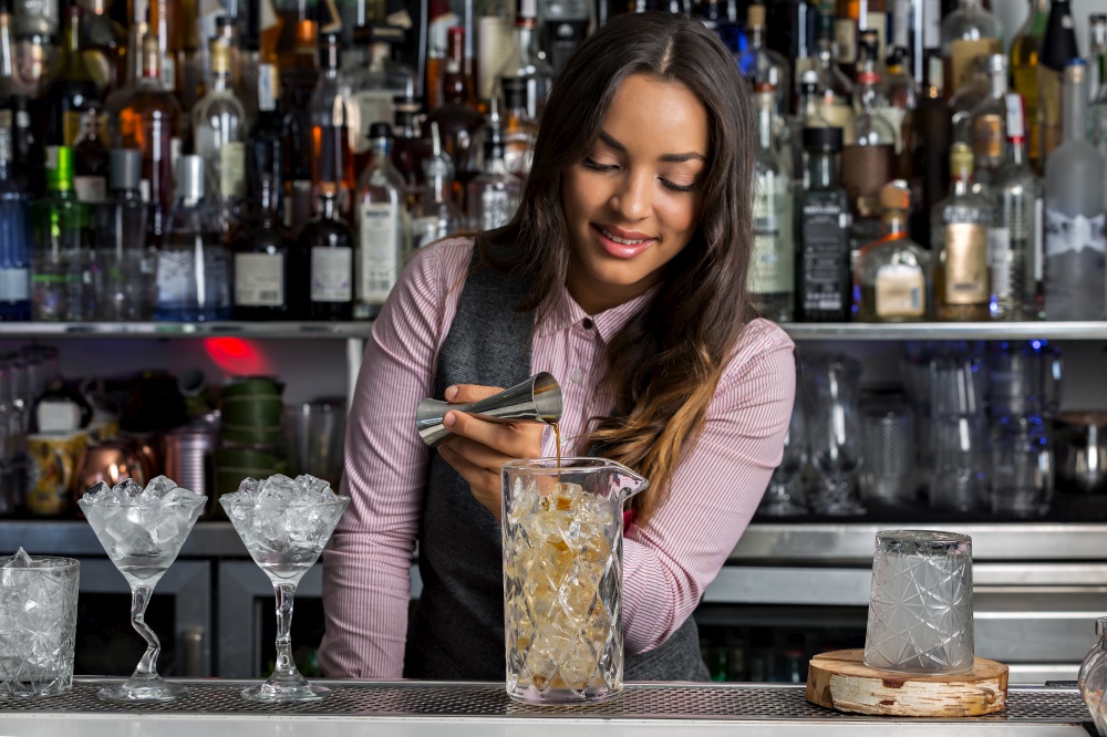 Young Hispanic female bartender adding alcohol from stainless jigger into mixing glass with ice cubes while preparing cocktail at bar counter. Positive female barkeeper making cocktail in bar