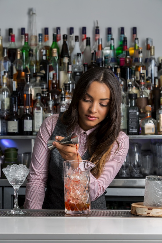 Young Hispanic female mixologist or bartender pouring alcohol drink from jigger into mixing glass while making cocktail at bar counter. Professional female mixologist preparing cocktail in bar
