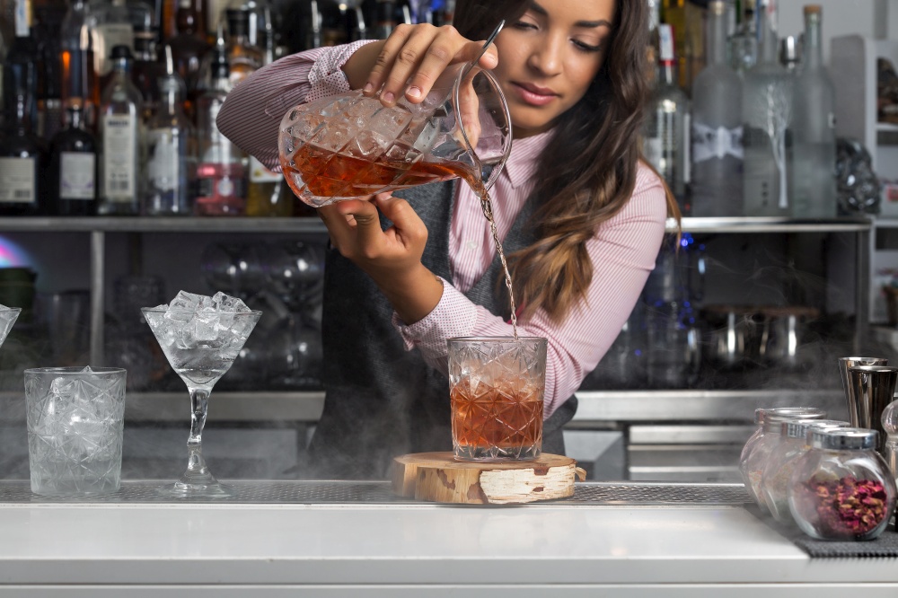 Skilled young ethnic female bartender pouring cocktail into glass while serving drink for order at bar counter. Woman preparing cocktail at bar counter