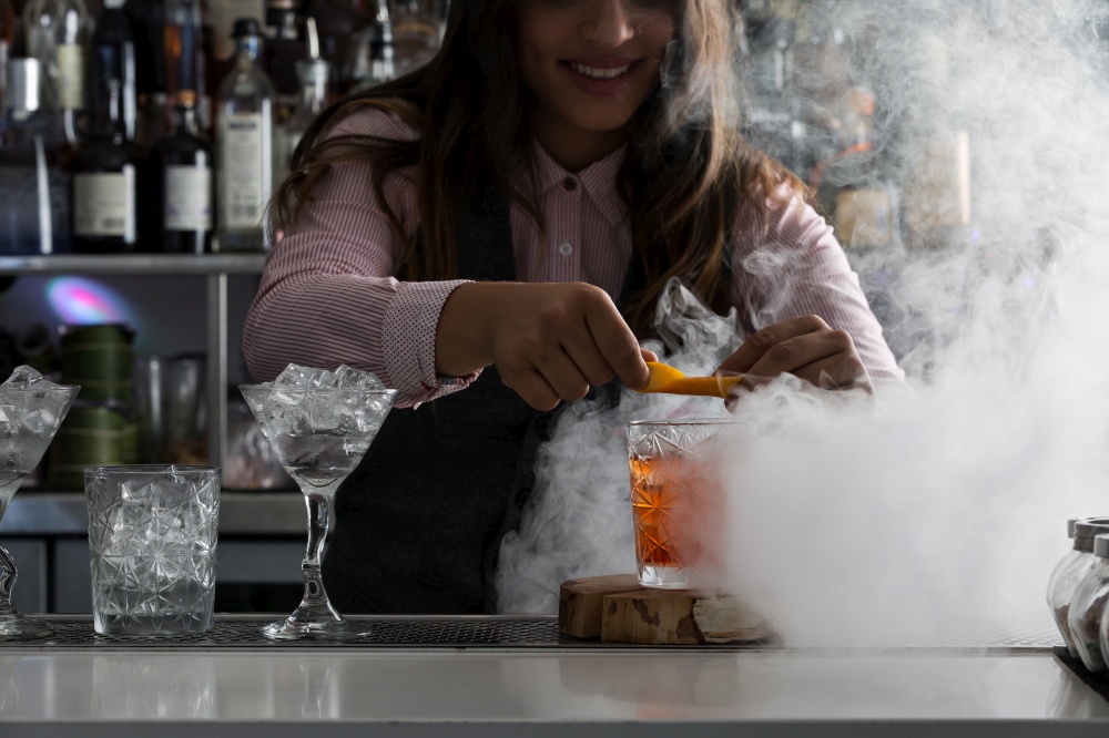 Crop smiling female barkeeper garnishing alcohol cocktail with orange slice while preparing drink at bar counter with steam. Bartender preparing cocktail at counter with steam