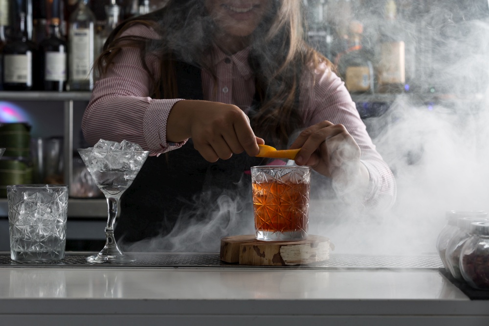 Crop cheerful female bartender adding fresh orange peel into glass with alcohol drink while working at bar counter with steam. Barkeeper serving cocktail at bar counter