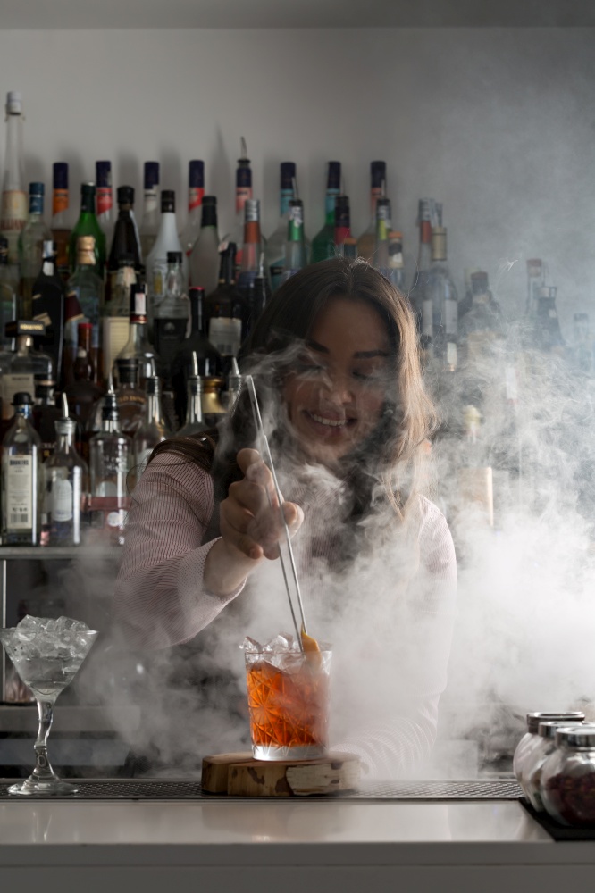 Positive young female barkeeper with tongs adding ingredient into glass with cold alcohol beverage with ice cubes while working at bar counter with steam. Cheerful bartender making cocktail in bar