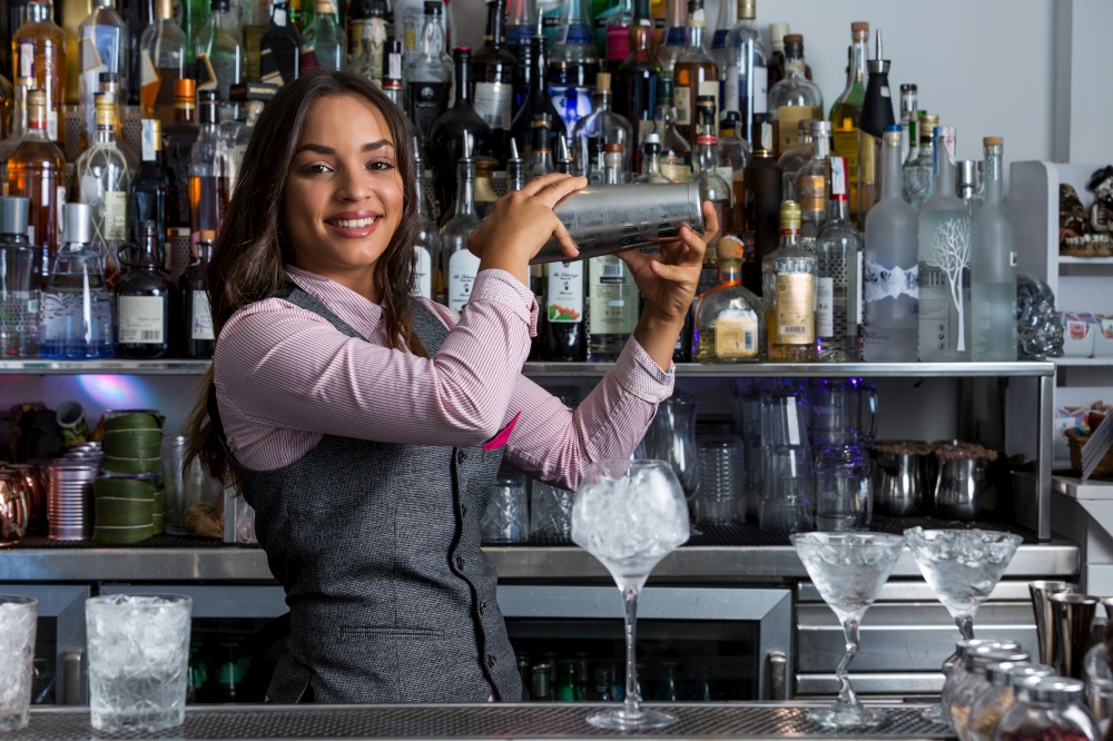 Happy young ethnic female barkeeper mixing drinks in shaker while preparing cocktail with ice cubes at bar counter. Cheerful female bartender with shaker preparing cocktail