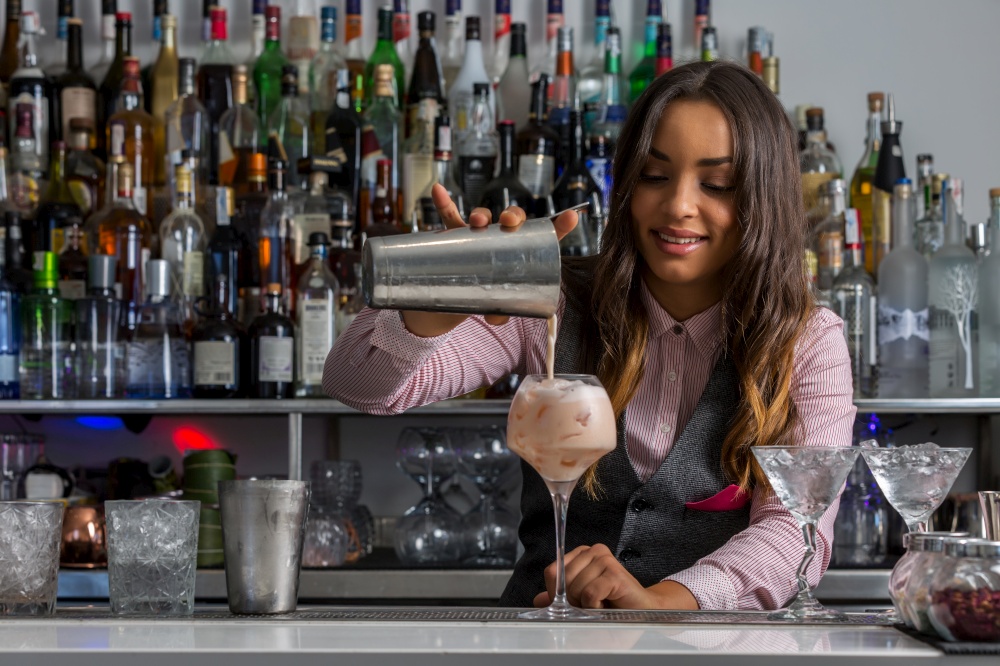 Smiling young Hispanic female bartender pouring foamy cocktail from shaker into glass goblet while serving drinks at bar counter. Female barkeeper pouring cocktail into glass