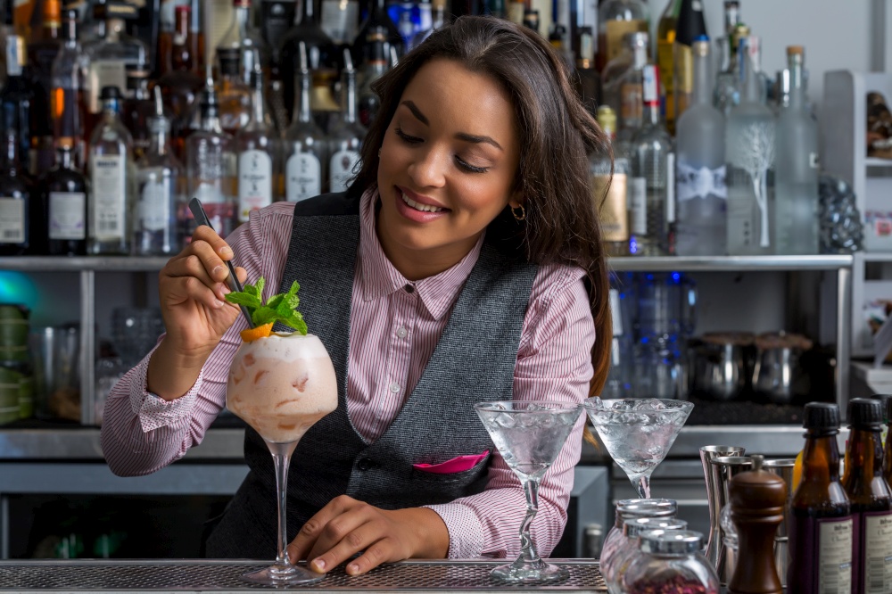 Cheerful Hispanic female mixologist decorating sweet cocktail with fresh mint and orange peel during work in pub. Happy Hispanic woman decorating cocktail in bar