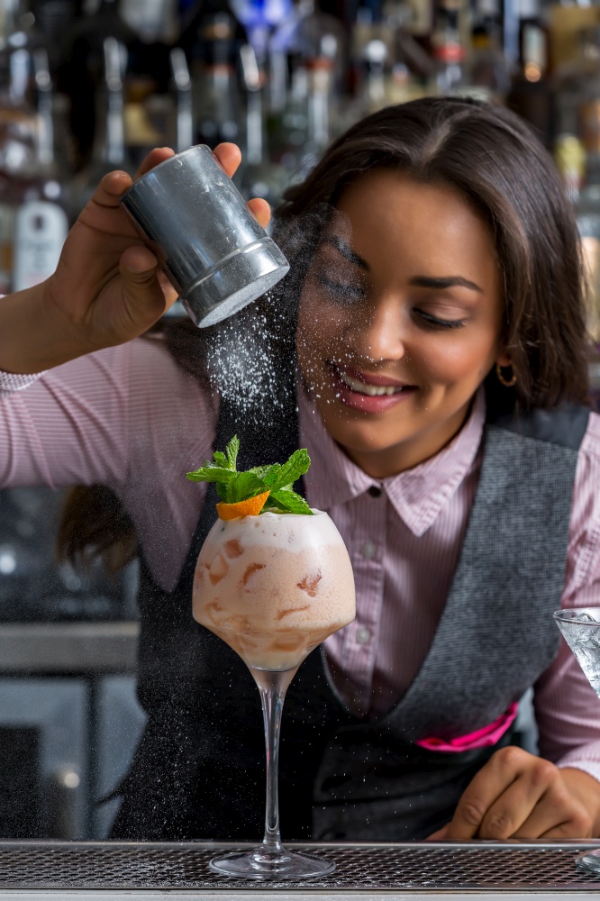 Glad young Hispanic woman smiling and adding powdered sugar to fresh cocktail during work in pub. Optimistic female bartender preparing sweet cocktail