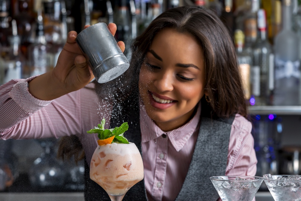 Hispanic woman smiling and adding icing sugar on top of sweet cocktail with mint and orange peel during work in bar. Happy female barkeeper adding powdered sugar to cocktail