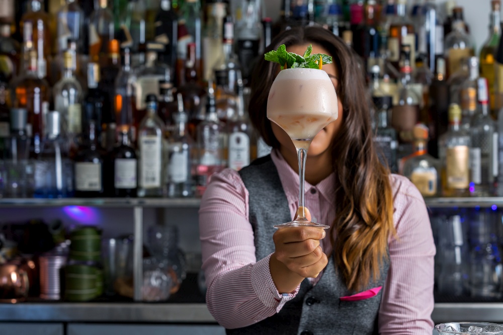 Anonymous woman demonstrating glass of fresh cocktail with mint leaves against shelf with alcohol in bar. Unrecognizable female mixologist showing cocktail