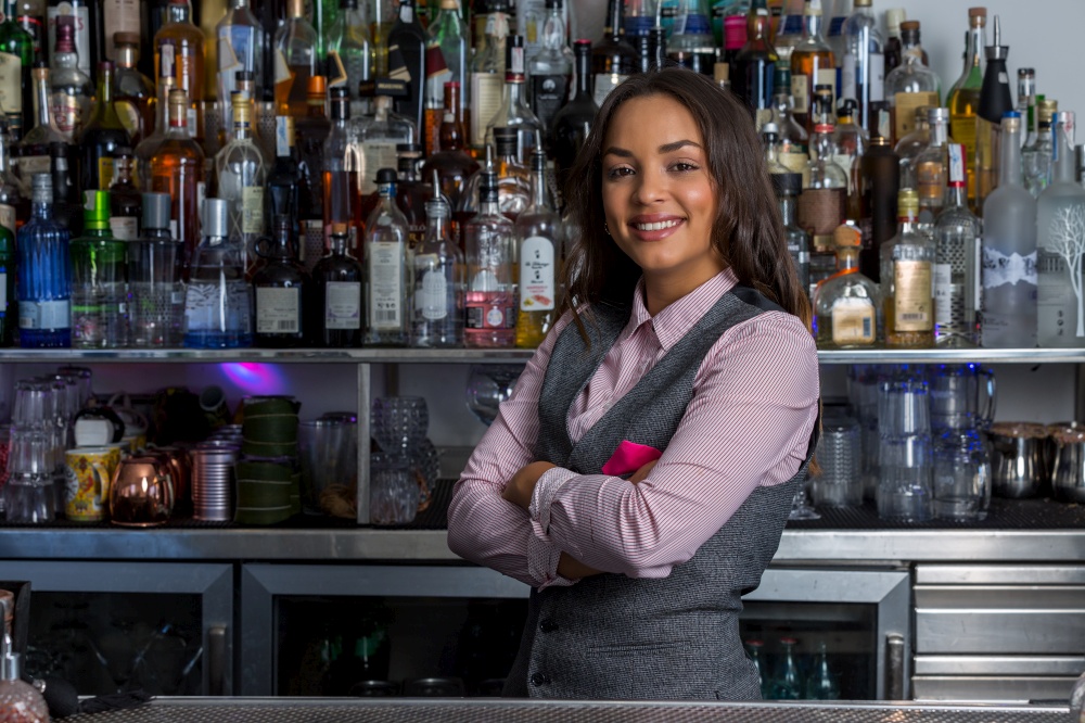 Positive Hispanic female bartender crossing arms and looking at camera with smile during work in pub. Confident Hispanic woman working in bar