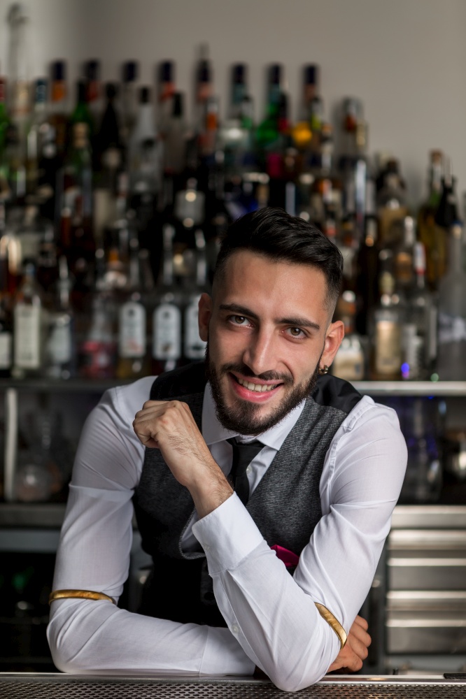 Smiling young bearded male bartender in elegant outfit leaning on bar counter and looking at camera against blurred bottles of alcoholic drinks. Handsome barkeeper working at counter