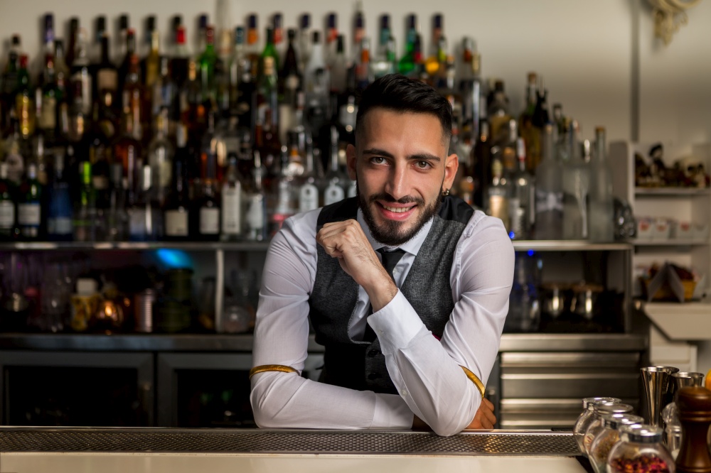 Handsome young bearded Hispanic male barkeeper in elegant clothes smiling friendly and looking at camera while standing behind bar counter with alcoholic drinks. Smiling barman standing at bar counter