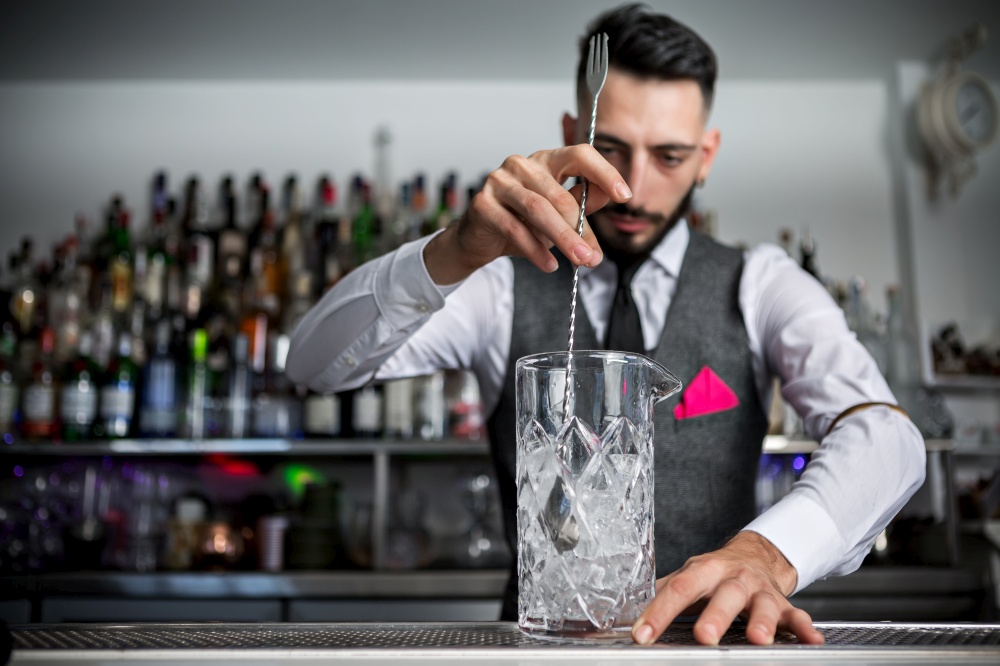 Professional barman stirring alcohol cocktail with long spoon in mixing glass while working at counter. Bartender mixing cocktail in glass