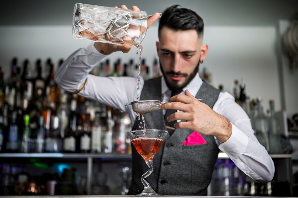 Skilled young Hispanic barman in elegant clothes filling goblet with alcohol cocktail through metal sieve while working at bar counter. Bartender with sieve pouring drink into glass