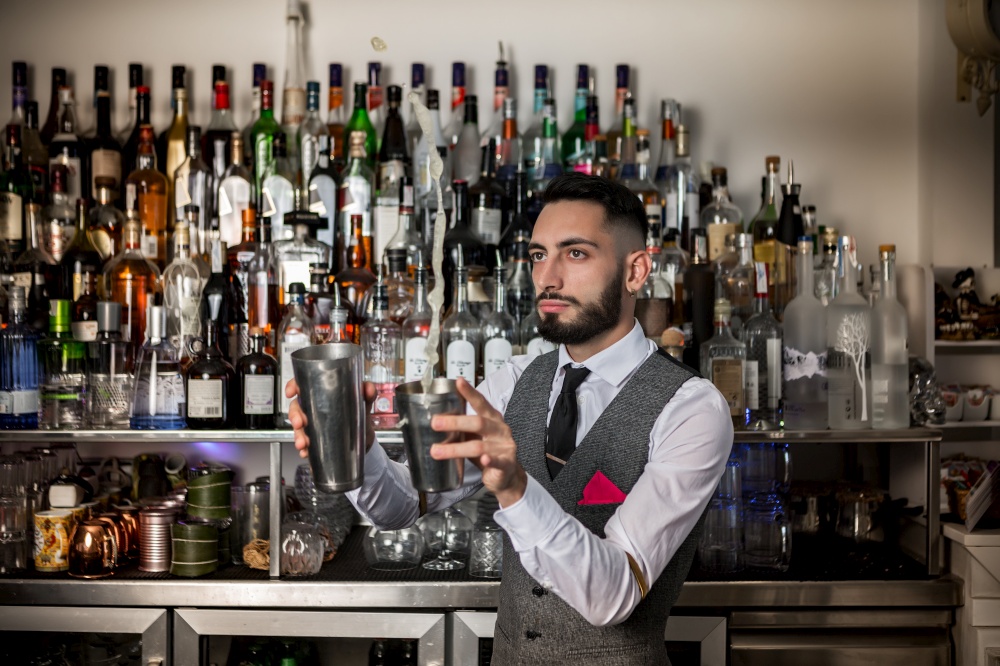 Experienced Hispanic male bartender with stainless shaker performing trick while mixing alcohol beverage and preparing cocktail in bar. Professional barman with shaker preparing alcoholic drink